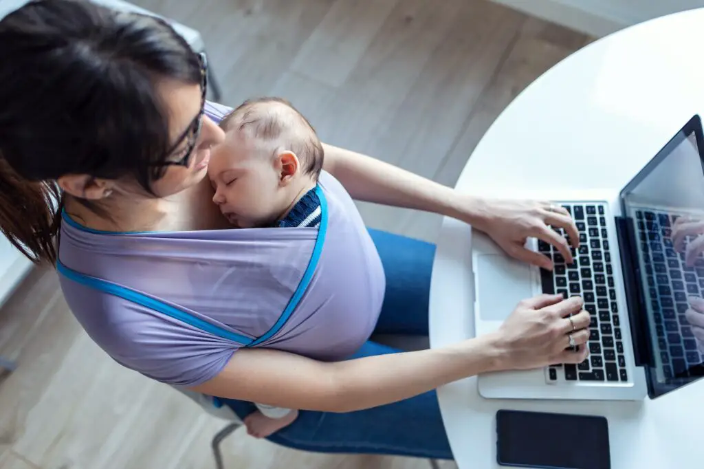 Pretty young mother with her baby in sling working with laptop at home.