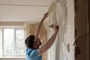 Hardworking man removes old wallpaper from wall with spatula in room. Home repairs.