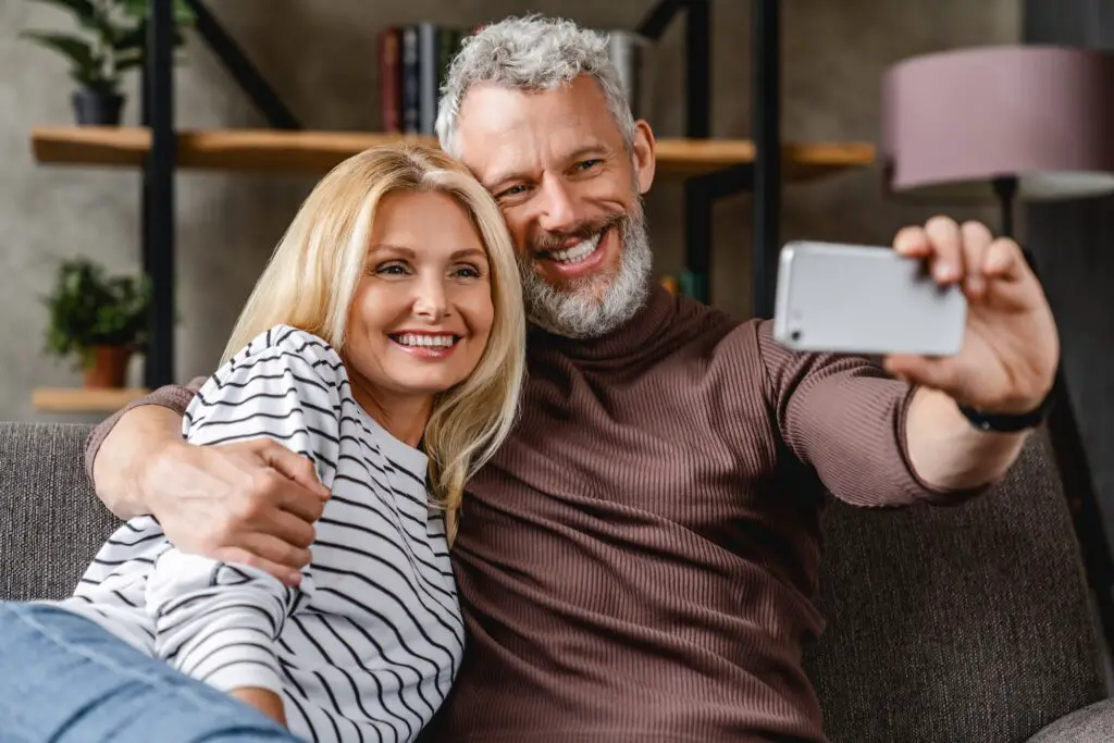 Modern middle aged couple sitting on sofa at home and making self-portrait on smartphone