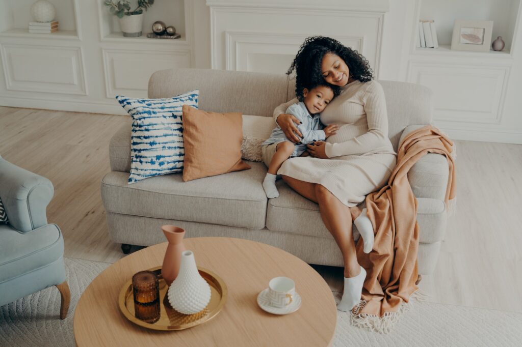Single african pregnant mom tenderly embracing little son while sitting on cozy couch